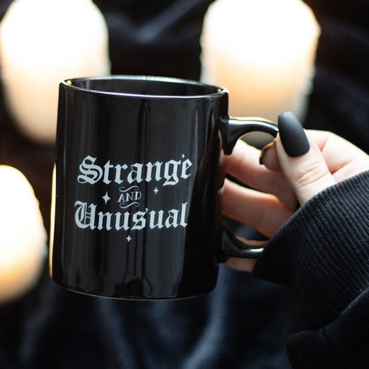 Black mug with 'Strange and Unusual' text held by a person against a blurred candlelit background