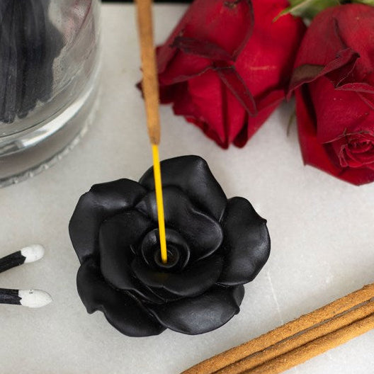 Black rose-shaped incense burner with a yellow incense stick, surrounded by red roses and wooden sticks on a white surface.