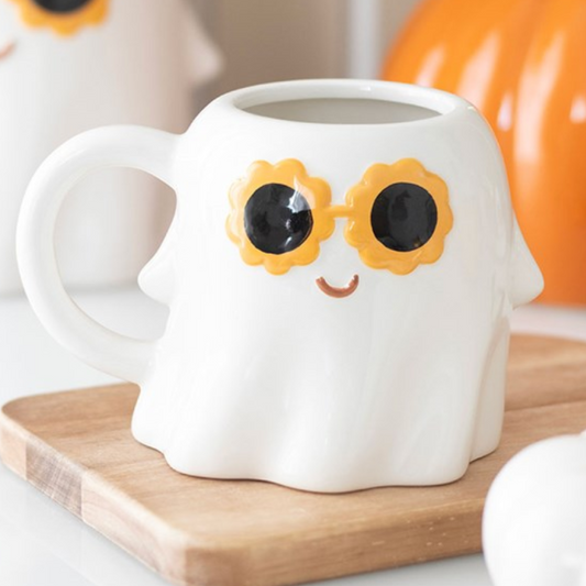 White ghost-shaped mug with sunflower sunglasses on a wooden coaster, with pumpkins in the background.
